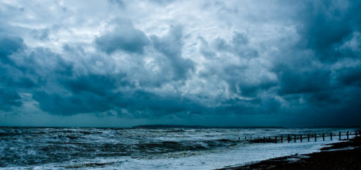 A windy day looking across the bay, with stormy blue-white clounds in the sky and rough waves breaking against a shingle beach 